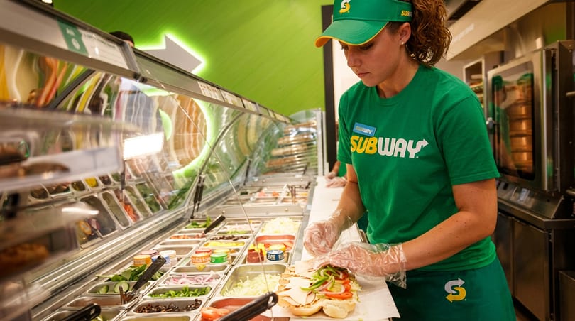 A Subway employee in a green uniform prepares a sandwich with fresh ingredients at the counter.