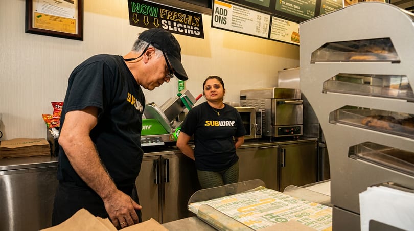 Two Subway employees behind a counter with a "NOW FRESHLY SLICING" sign.