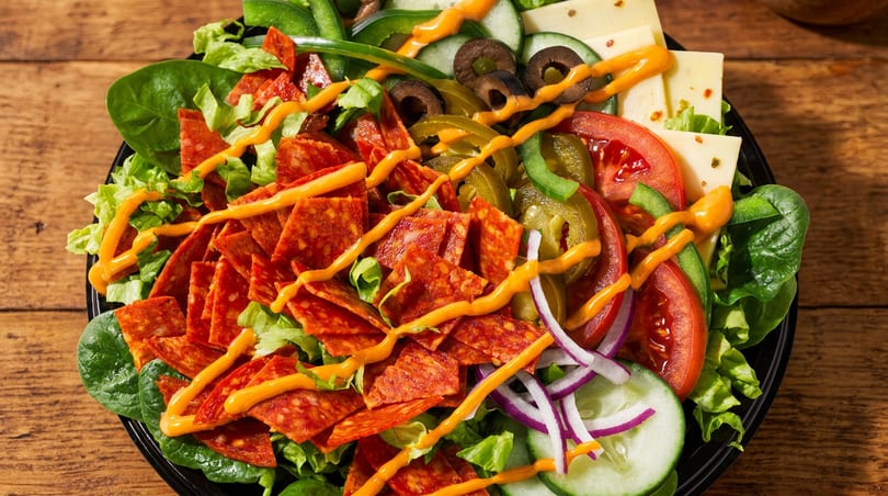 Pepperoni salad with mixed greens, sliced vegetables, cheese, and orange dressing in a black bowl.