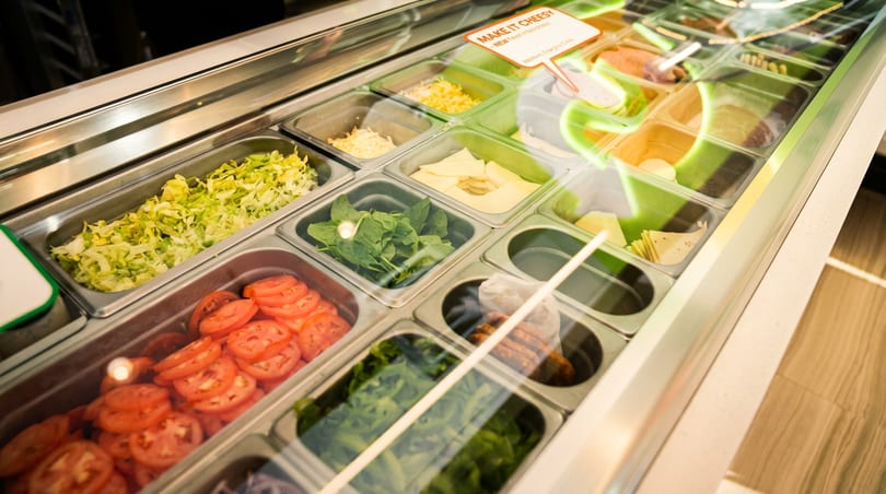 Refrigerated food display counter with fresh lettuce, tomatoes, spinach, and cheese in various bins.