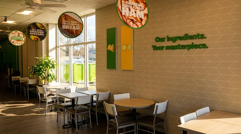 Sunny Subway dining area with tables, chairs, ingredient signs, and a 'Your masterpiece' wall quote.